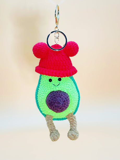 Crocheted avocado keychain with a red hat on a plain background
