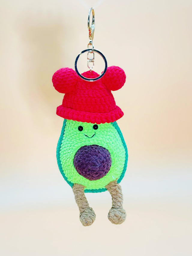 Crocheted avocado keychain with a red hat on a plain background